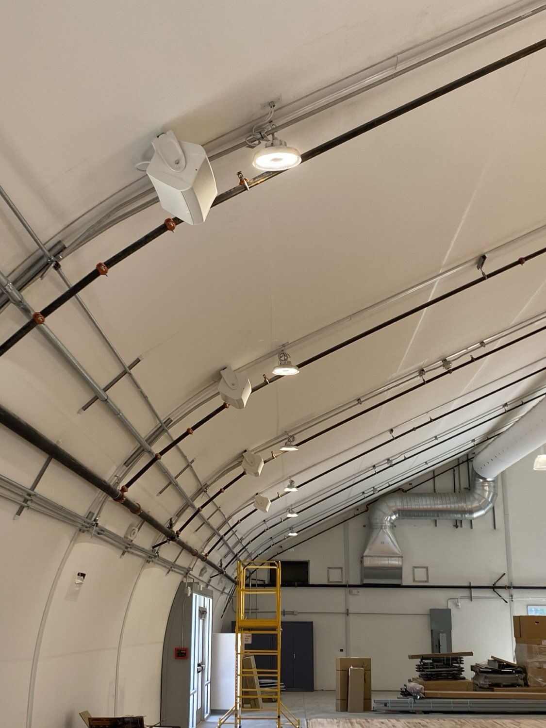 Commercial ceiling-mounted white speakers wired in sequence along the curved structural ribs of an athletic facility