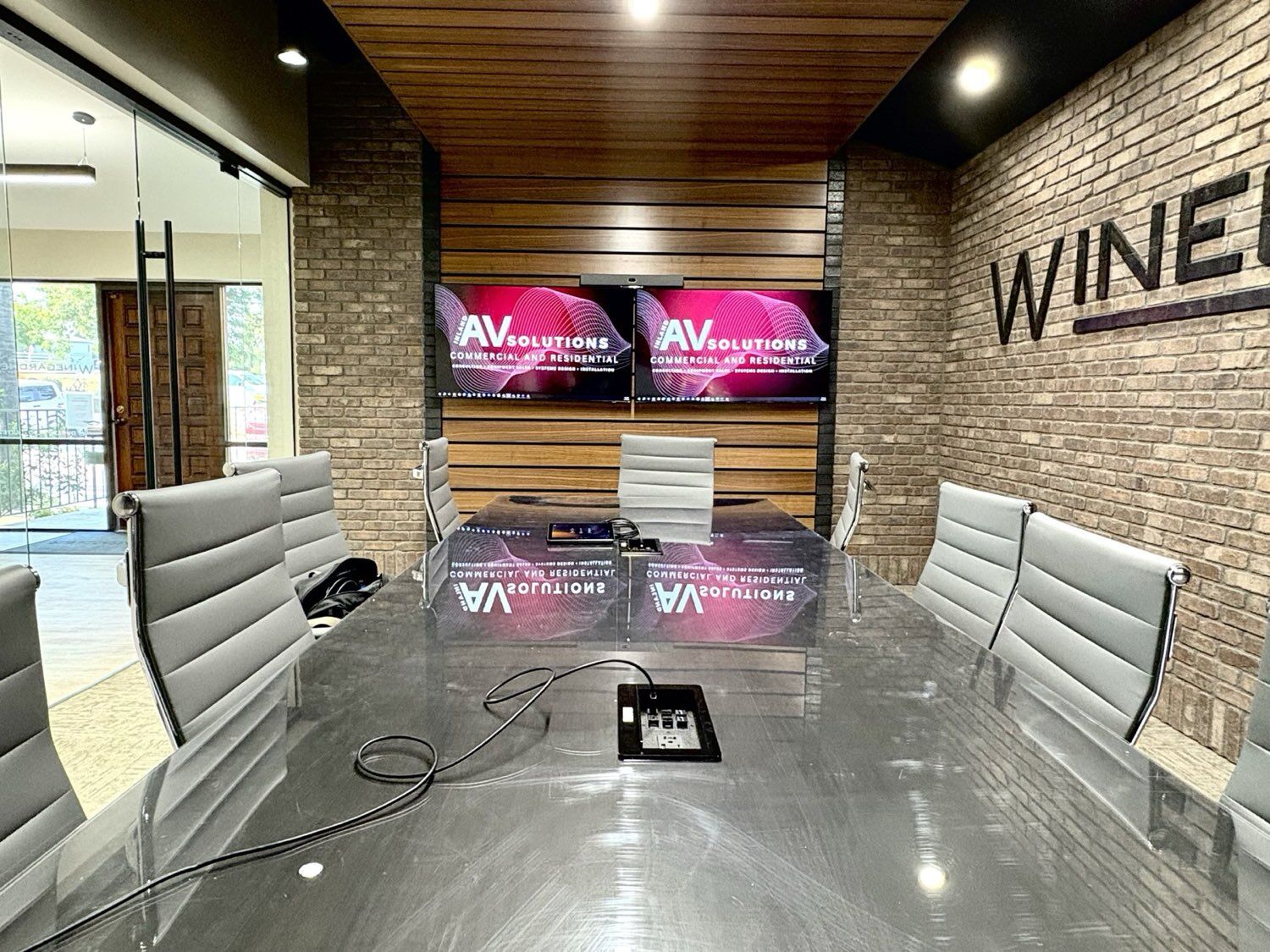 Finished executive boardroom with exposed brick, wood-slat accent wall, glass conference table, and dual displays showing Inland AV Solutions branding