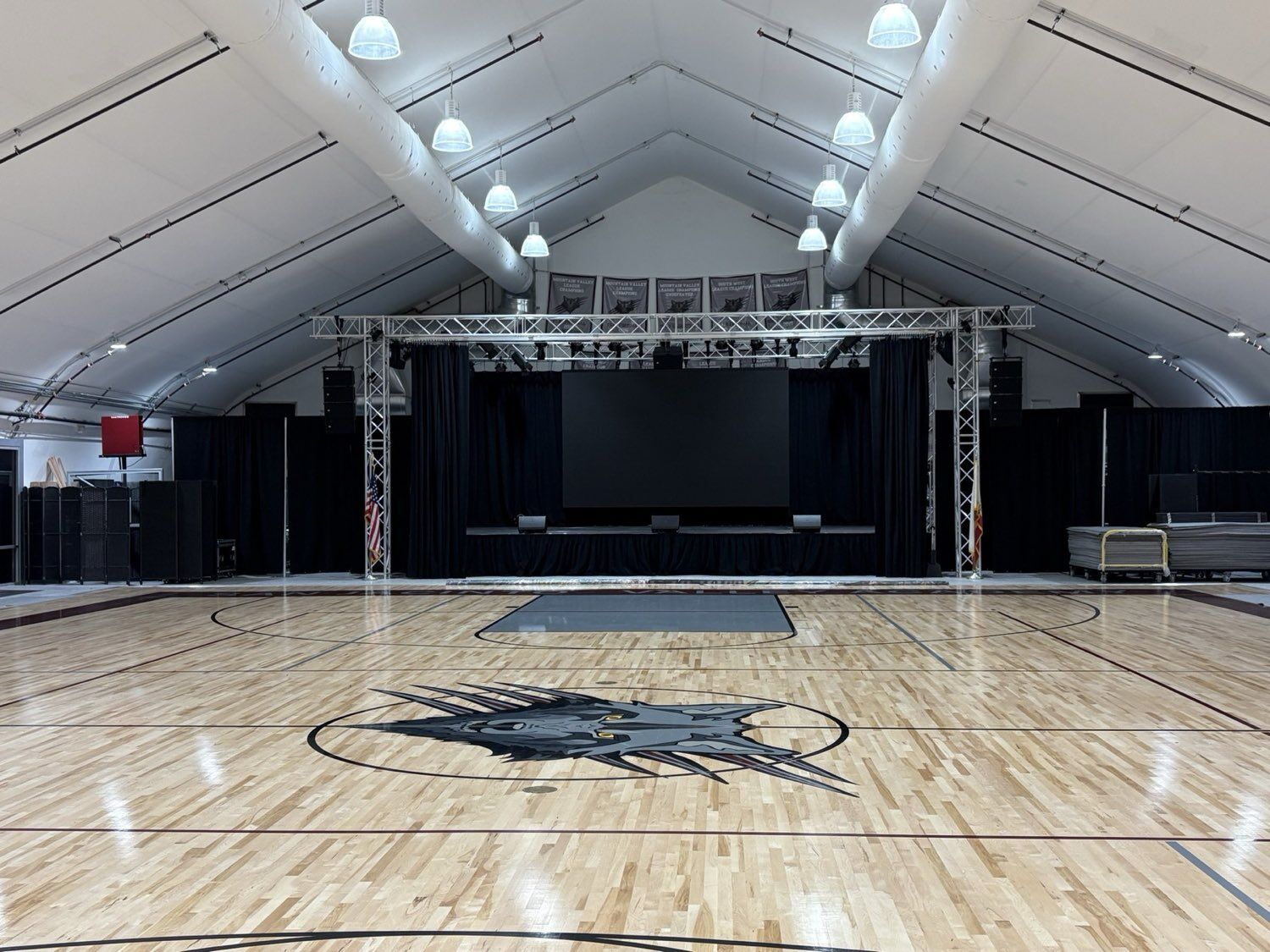 Wide view of the SJVA athletic facility, full basketball court, championship banners, and a truss-mounted stage with projection screen