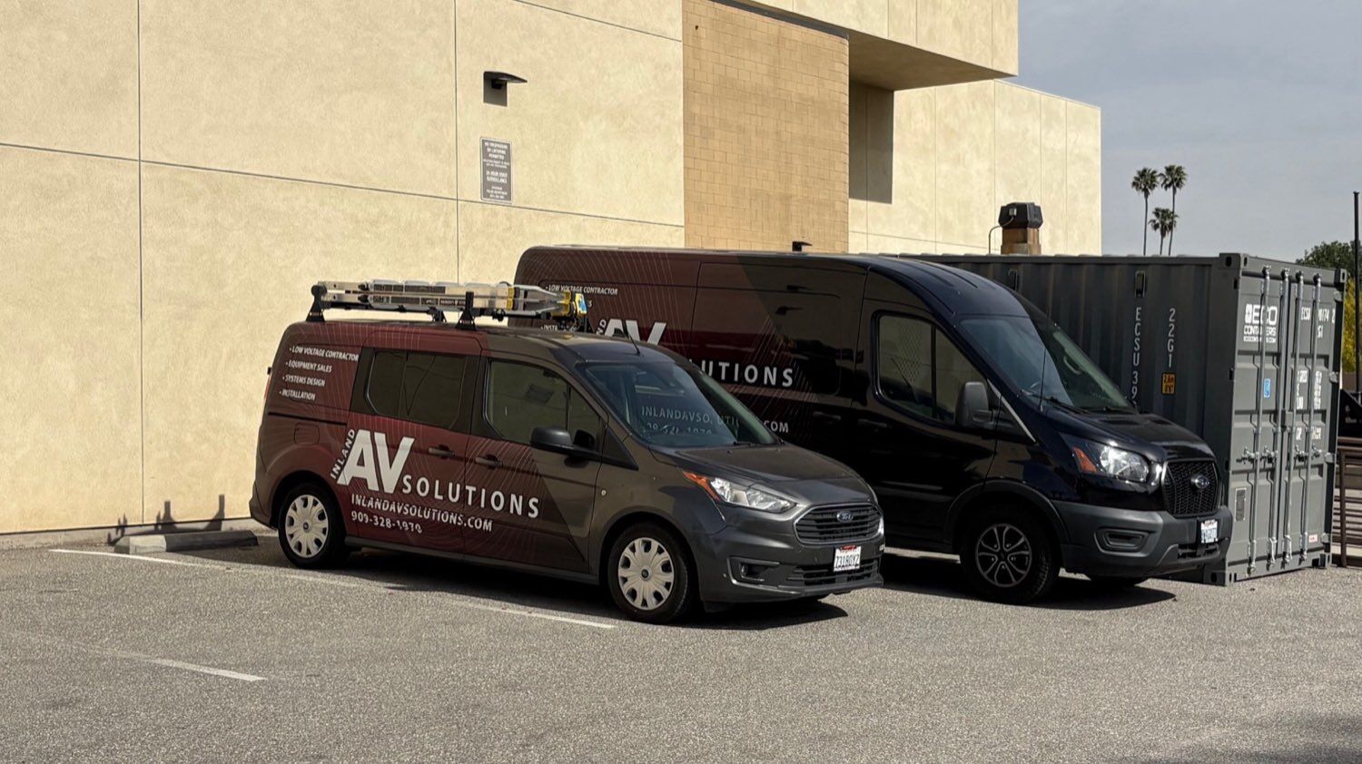The Inland AV Solutions company fleet, two charcoal and maroon branded service vans parked beside a commercial building under Southern California palm trees